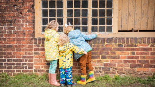 Three young children wearing colourful raincoats and wellies peer through the window of one of the stableyards buildings as part of the Easter trail at Calke Abbey.